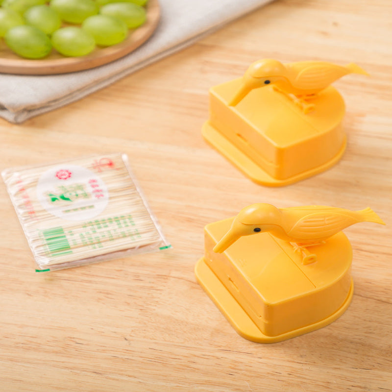 Two yellow bird-shaped containers on a wooden surface with a pack of toothpicks and grapes in the background.