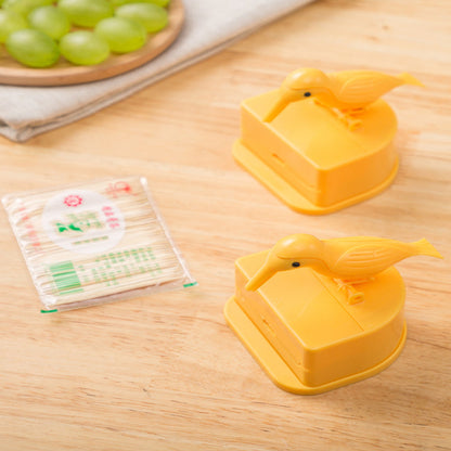 Two yellow bird-shaped containers on a wooden surface with a pack of toothpicks and grapes in the background.