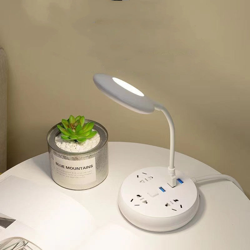 Desk lamp with power strip on a white surface next to a potted plant.