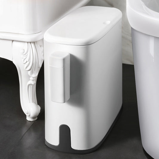 White trash can with foot pedal on a dark floor next to a white toilet.